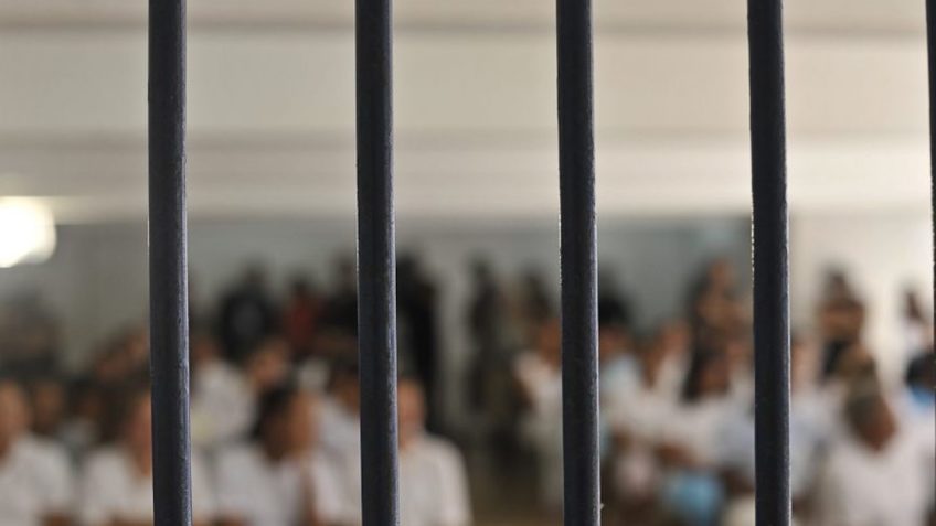Grupo de pessoas, sentadas e em pé, em uma cela em um estabelecimento carcerário