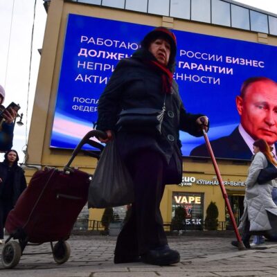 Pessoas passam diante de monitor com a imagem e uma citação de Putin em São Petersburgo