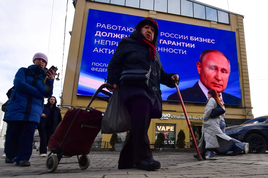 Pessoas passam diante de monitor com a imagem e uma citação de Putin em São Petersburgo