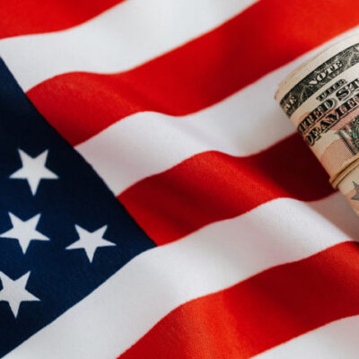 Foto colorida horizontal. Cédulas de dinheiro enroladas com elástico sobre uma bandeira dos Estados Unidos.
