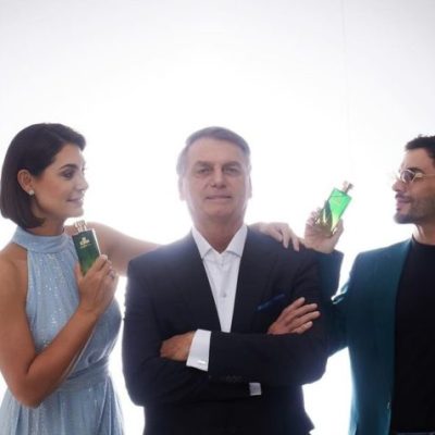 Michelle Bolsonaro, de vestido azul, e Agustin Fernandez, de terno verde, seguram o perfume de Jair Bolsonaro. O ex-presidente está entre os dois sentado em um banco com os braços cruzados