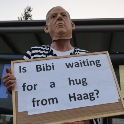 Ativista de esquerda usa máscara do premier de Israel, Benjamin Netanyahu, e roupa de presidiário, e ergue cartaz com dizeres: 'Bibi está esperando por um abraço de Haia?'