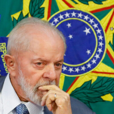 O presidente Luiz Inácio Lula da Silva com a mão na boca e o brasão do Brasil atrás