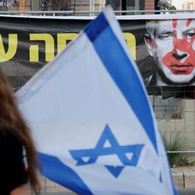 Mulher segura bandeira israelense enquanto passa por cartaz que representa o primeiro-ministro israelense  Benjamin Netanyahu com uma impressão de uma mão ensanguentada no rosto.