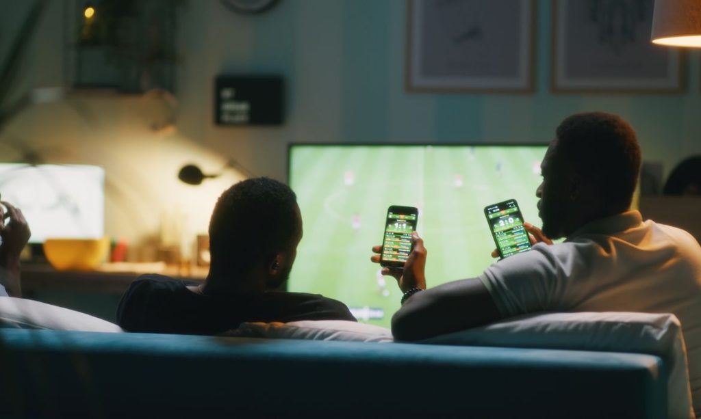 13/07/2026 - Bets - African American man using smartphone and clinking bottles of beer with friends while chilling on sofa and watching soccer game on TV. Foto: Framestock/ Adobe Stock