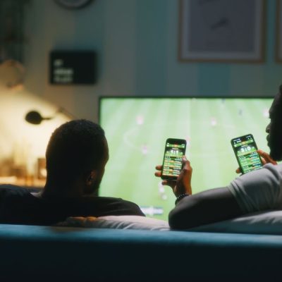 13/07/2026 - Bets - African American man using smartphone and clinking bottles of beer with friends while chilling on sofa and watching soccer game on TV. Foto: Framestock/ Adobe Stock