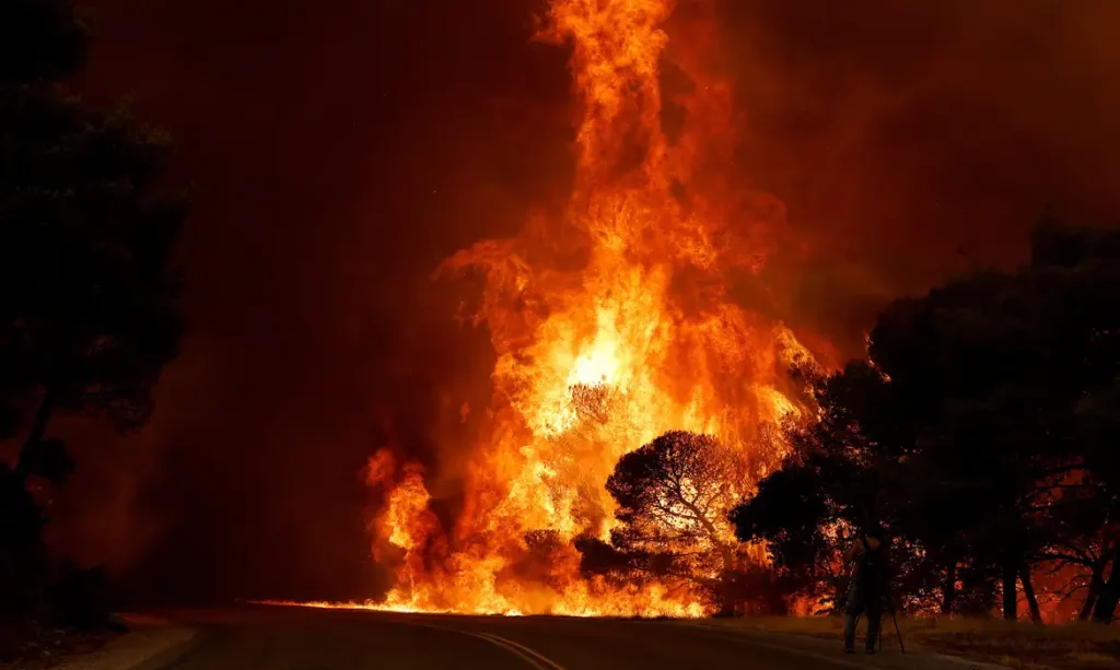 Um homem grava imagens enquanto a fumaça e as chamas se elevam de um incêndio florestal próximo a Agia Paraskevi, na Grécia, em 1º de agosto de 2026. REUTERS/Louisa Gouliamaki/Proibida Reprodução