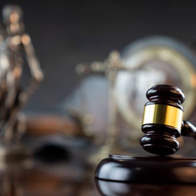 martelo de juiz em madeira escura e detalhes dourados se destaca em primeiro plano, tendo ao fundo, de forma desfocada, símbolos da justiça como a estátua da Temis e uma ampulheta