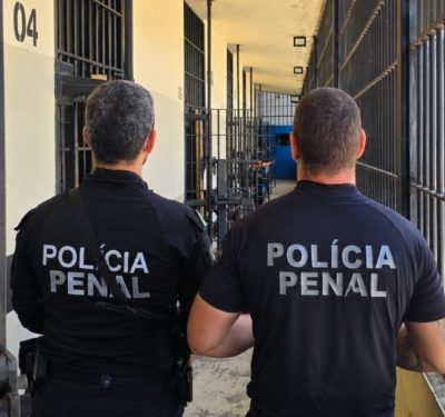 Dois policiais penais de costas em um corredor de um presídio em frente a celas com grades de ferro.