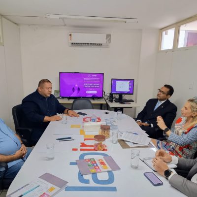 Grupo de pessoas reunido ao redor de uma mesa de reunião em uma sala de escritório, enquanto conversam diante de uma tela ao fundo com uma mensagem roxa de conscientização.
