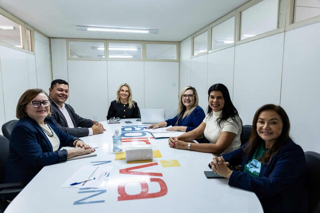 Seis pessoas estão reunidas e sorrindo ao redor de uma grande mesa branca de escritório em uma sala de reuniões iluminada.