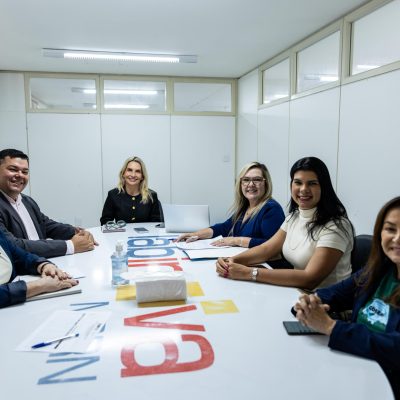 Seis pessoas estão reunidas e sorrindo ao redor de uma grande mesa branca de escritório em uma sala de reuniões iluminada.