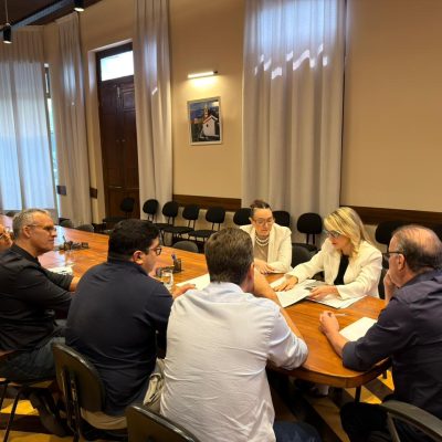 seis pessoas sentadas em mesa de reuniao com o prefeito a ponta e promotoras ao lado