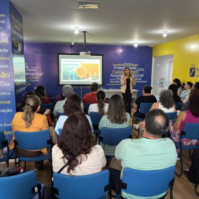 Uma mulher em pé fala ao microfone diante de uma plateia sentada em um auditório colorido, assistindo a uma apresentação de slides.