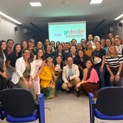 Grupo de pessoas sorrindo posa para foto em um auditório, reunidas à frente de uma tela com a apresentação do "Projeto Proteção Integrada".