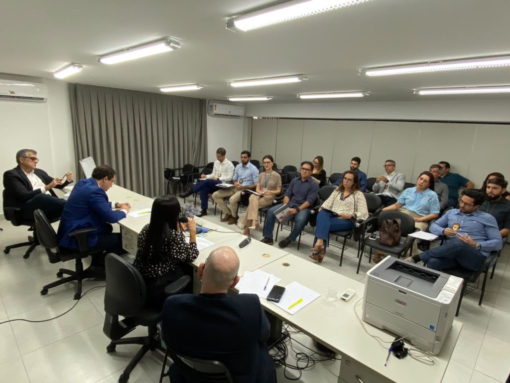 Grupo de pessoas reunido em uma sala bem iluminada durante uma reunião de trabalho em torno de uma grande mesa.