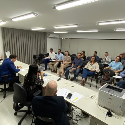 Grupo de pessoas reunido em uma sala bem iluminada durante uma reunião de trabalho em torno de uma grande mesa.