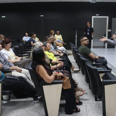 Uma mulher em terno cinza segura um microfone e fala para uma plateia sentada em um auditório.